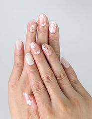 Hand model wearing short pink oval press-on nails with white floral designs, pearl details, and soft shimmer finish