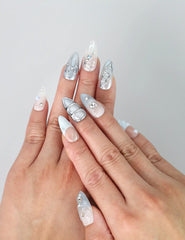 Hand model wearing blue almond-shaped press-on nails with iridescent pearl shimmer and sparkling 3D crystals