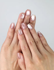 Hand model wearing short pink square press-on nails with white flower nail art, gold beads, and pearl embellishments
