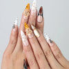 Hand model showing stiletto-shaped press-on nails in warm earth tones with bold tiger stripe patterns and floral 3D accents