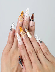 Hand model showing stiletto-shaped press-on nails in warm earth tones with bold tiger stripe patterns and floral 3D accents