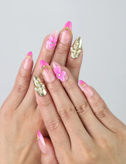 Hand model wearing handmade pink press-on nails with gold shimmer, 3D pink flowers, pearl accents, and tiny stars
