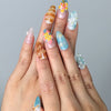 Hand model wearing almond-shaped press-on nails with pink floral art, starfish embellishments, and seashell-inspired texture