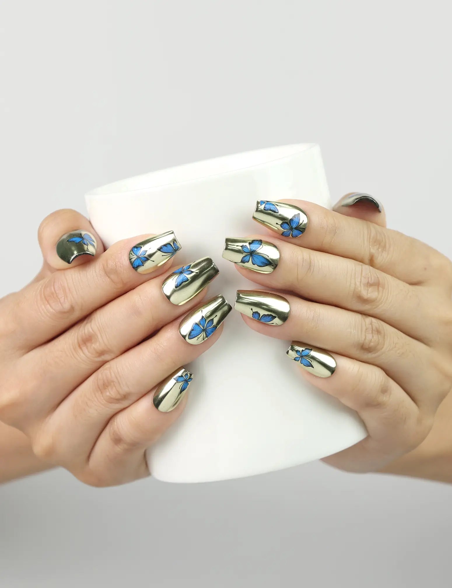 Hand holding a cup while wearing Blue Aura Singapore press on nails by naillover, highlighting golden sheen and butterfly details.