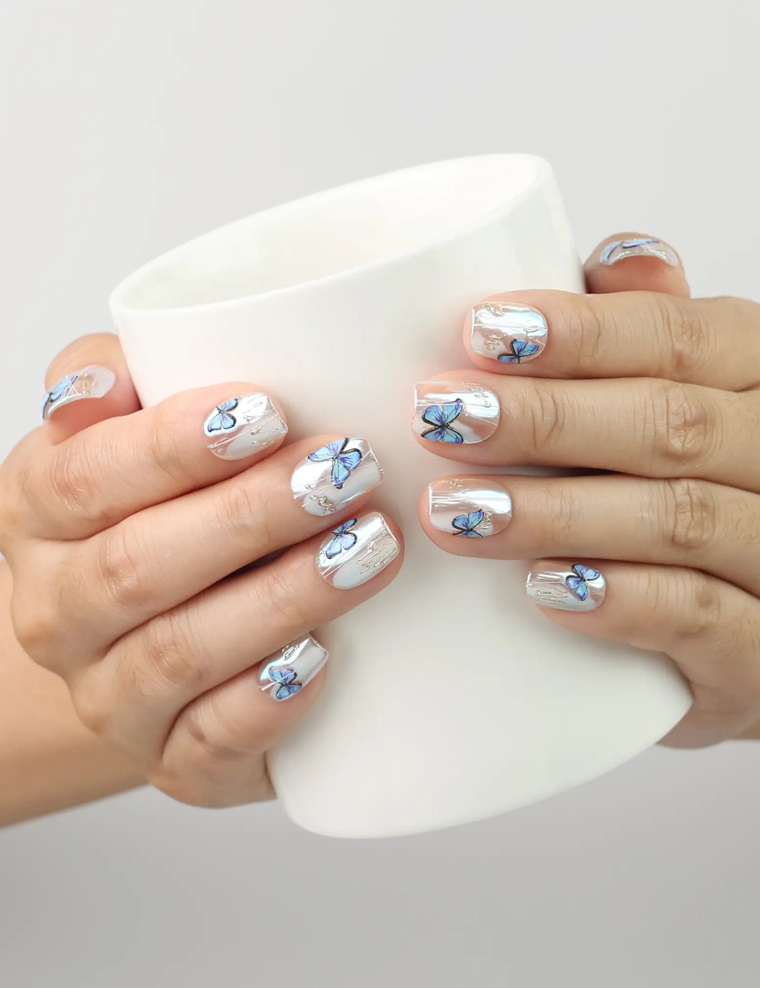 Hand holding a cup while wearing Butterfly Light short press-on nails with blue butterfly details from Naillover Singapore.