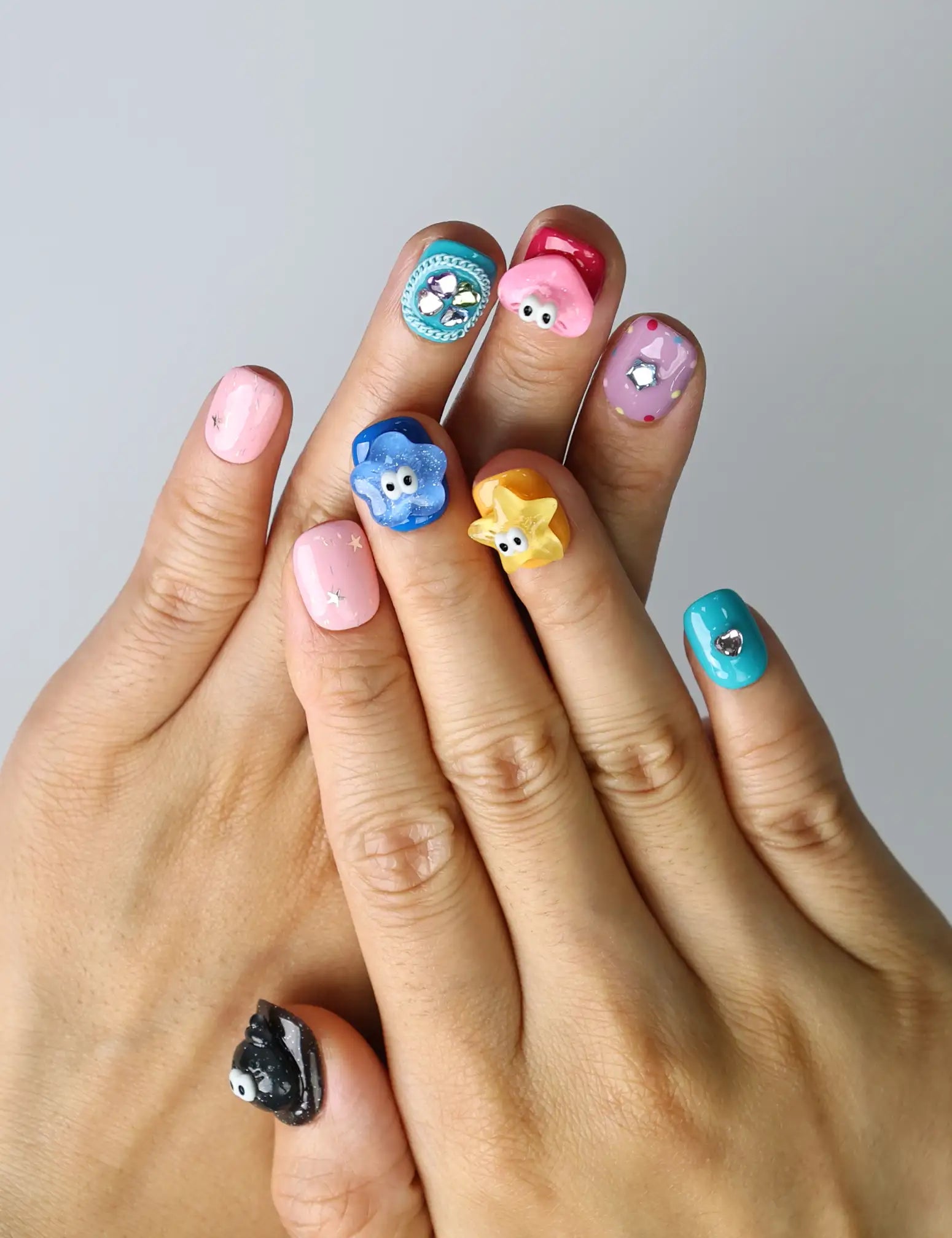 Hand model wearing short squoval press-on nails with playful 3D flowers, hearts, and multicolor googly eyes