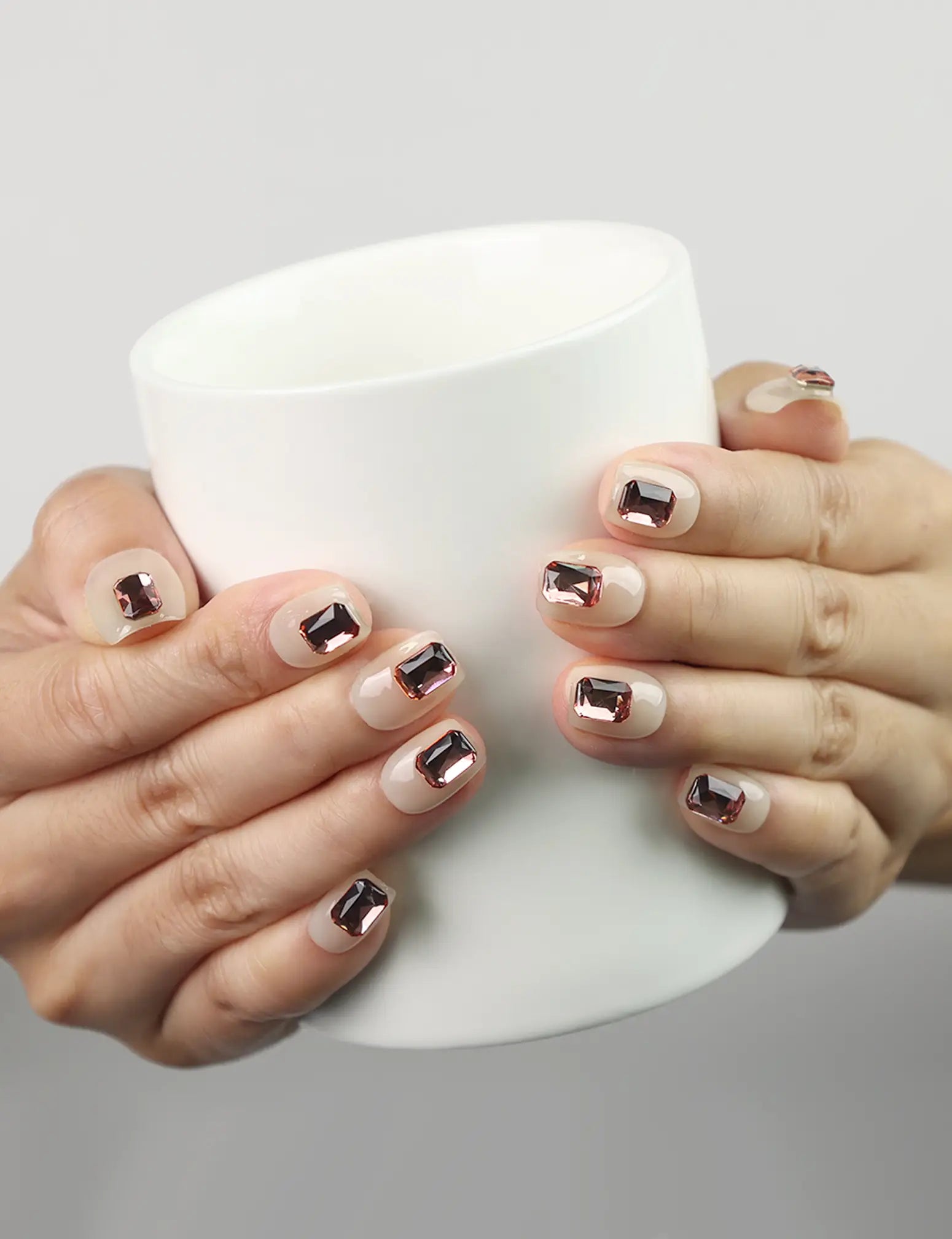 Hand holding coffee cup wearing Dream Glitter beige press-on nails with pink rhinestones, naillover trendy nails