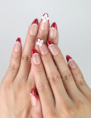 Hand model wearing almond-shaped red press-on nails with soft pink base, pearl details, and 3D floral embellishments