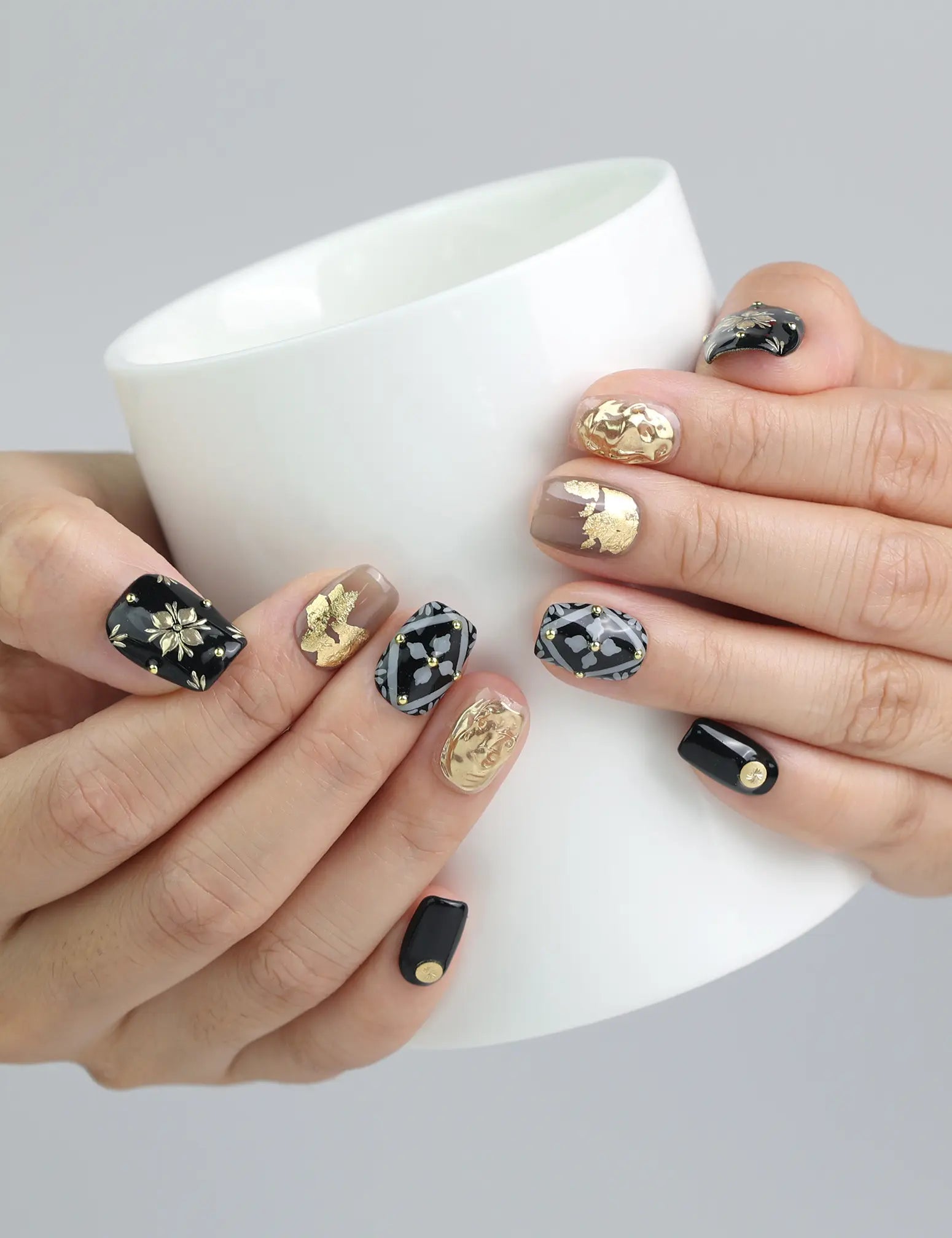 Hand holding a cup while wearing black handmade nails with white and gold floral designs, embellished with rhinestones by Naillover