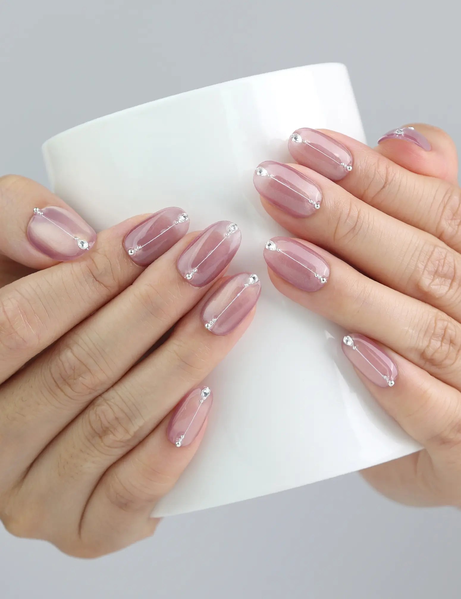 Hand holding a cup, showing Naillover’s mauve-pink gradient press-on nails with chrome shimmer and gemstones