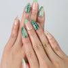 Hand model wearing nude and green short almond nails with gold foil and detailed tropical leaf nail art