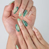 Close-up of handmade press-on nails in earthy nude and sage tones with palm leaf designs and shimmering gold accents