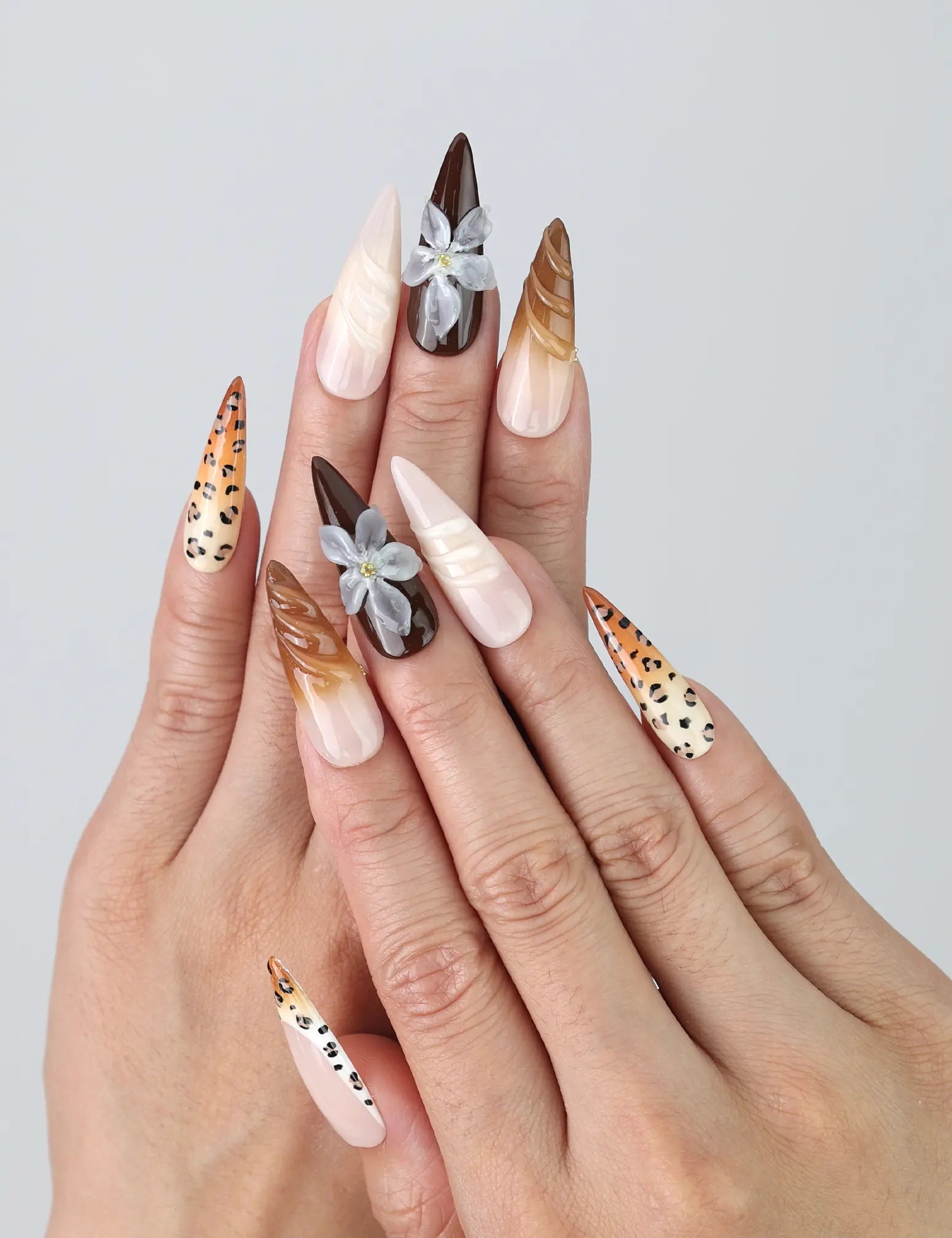 Hand model showing stiletto nails with bold brown leopard print, matte black accents, and delicate floral designs