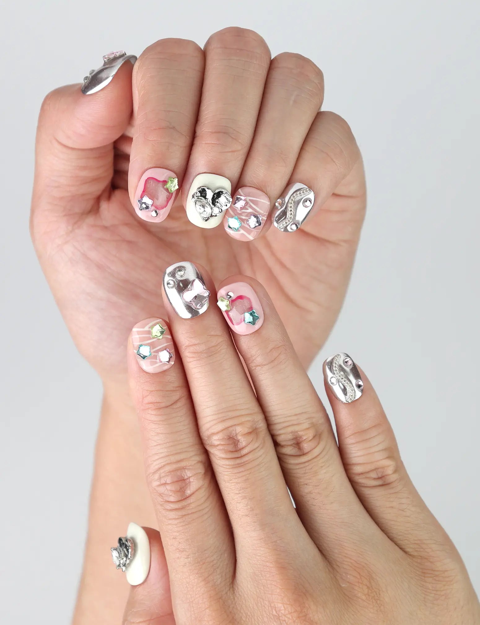 Hand model showing short silver-pink press-on nails with 3D hearts, pastel stars, and colorful gem decorations
