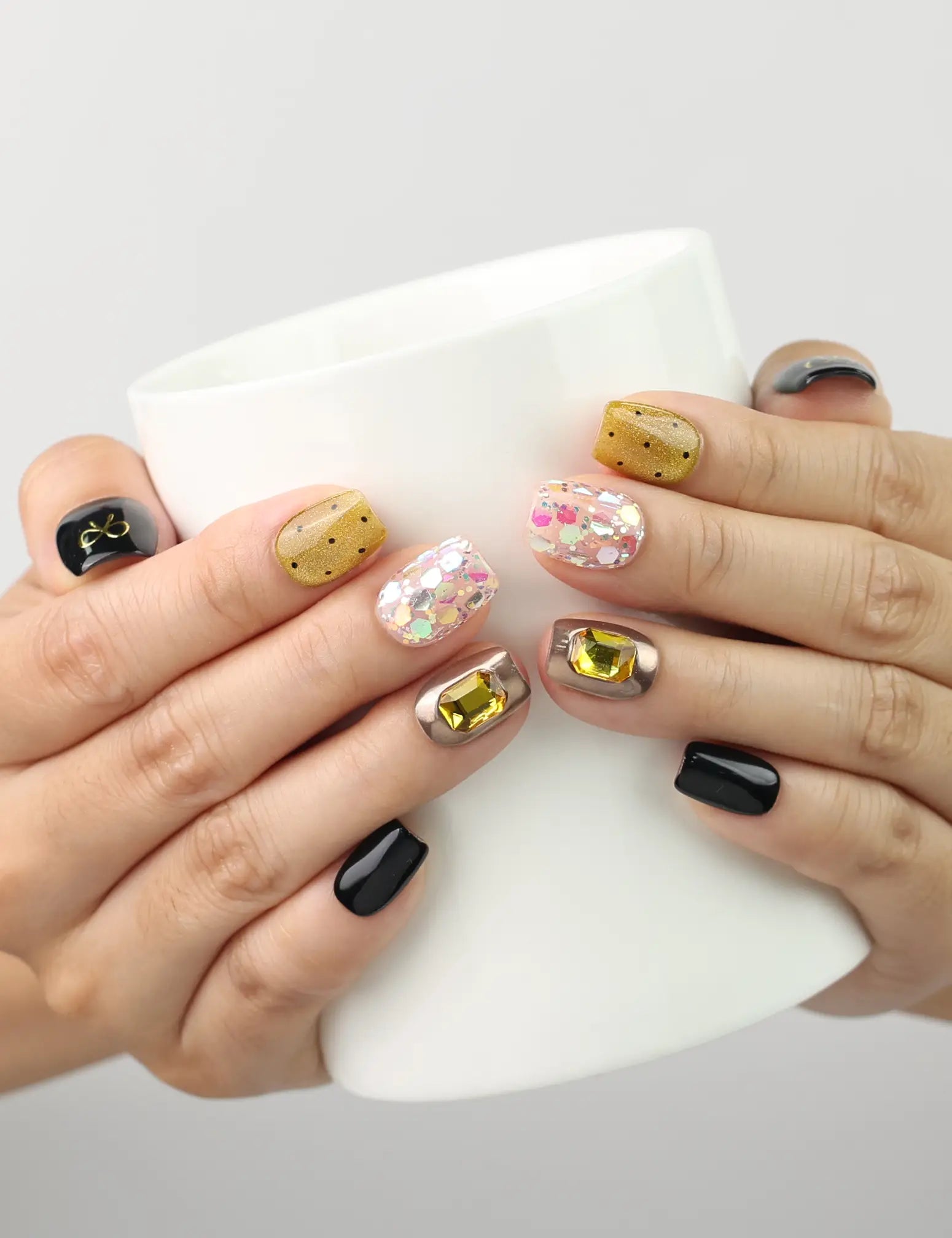 Hand holding a cup wearing Luxe Mix Singapore press on nails in black, gold, glitter, and gem accents, naillover