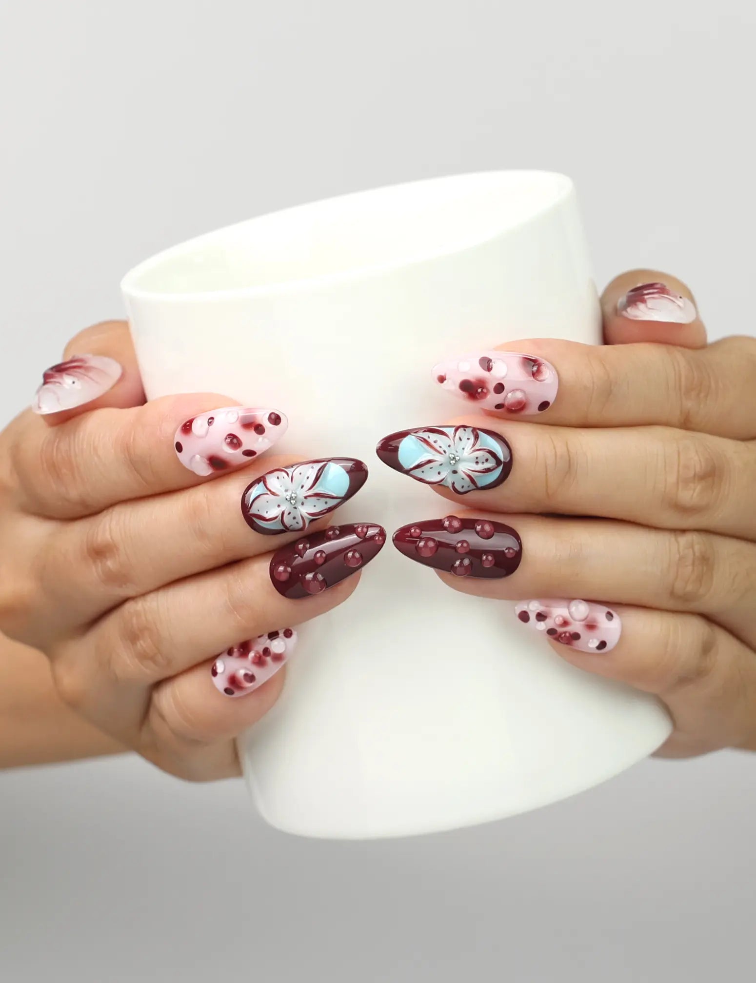 Model holding a cup while wearing Naillover Blush Enigma handmade press on nails with burgundy, white, floral, and polka dot design.