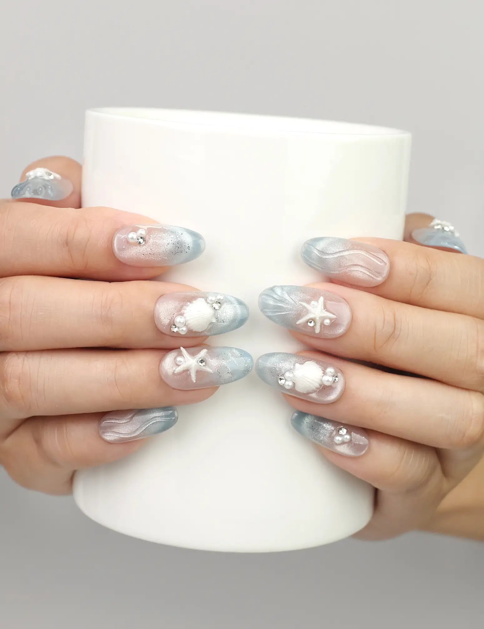 Hand holding a cup while wearing Naillover Ocean Whisper Singapore press on nails with blue gradient, pearls, and 3D starfish design