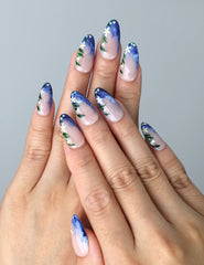 Hand model wearing blue almond-shaped press-on nails with hand-painted wave textures and soft floral accents