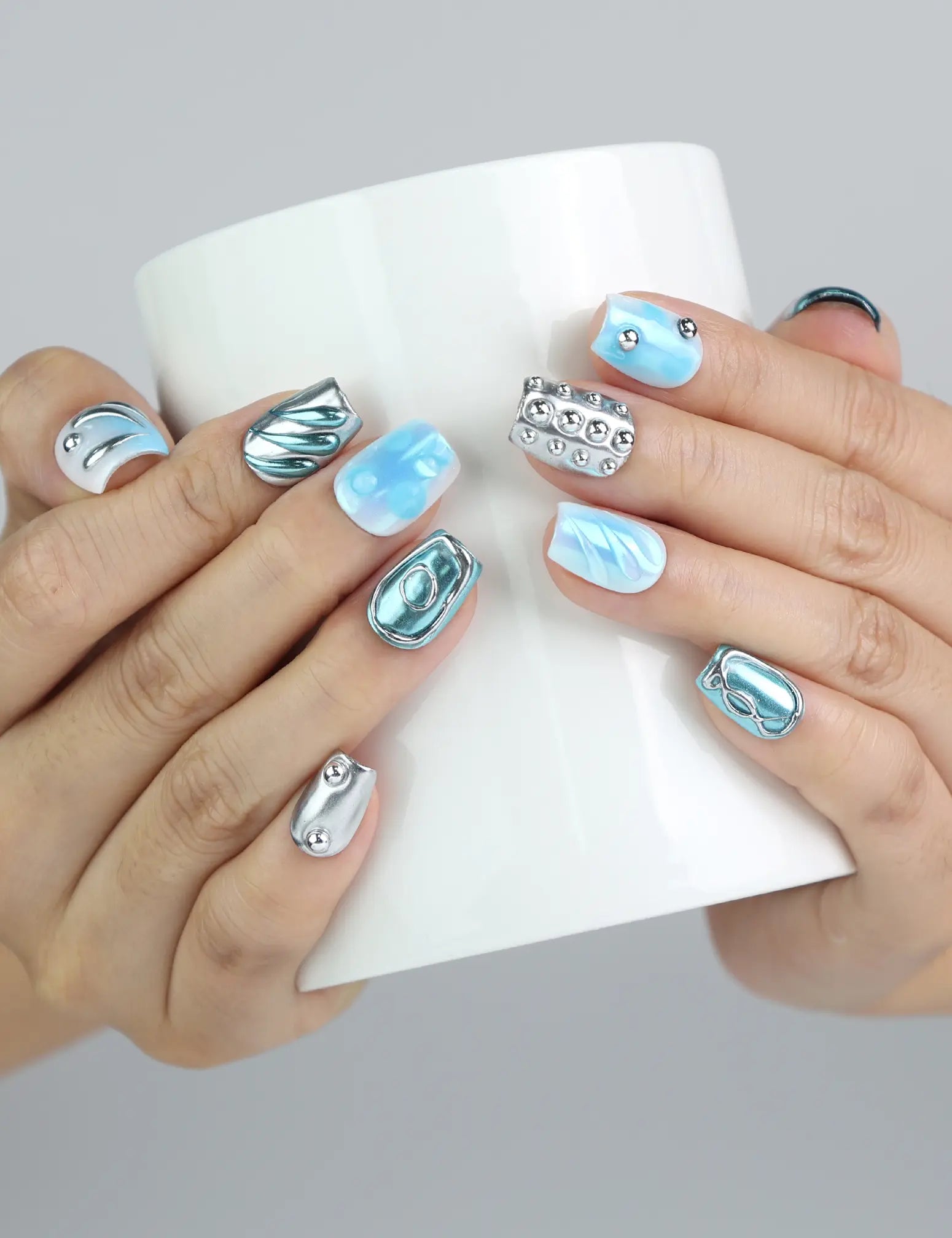 Hand holding a cup, displaying Naillover’s blue short coffin press-on nails with water droplet texture and metallic accents