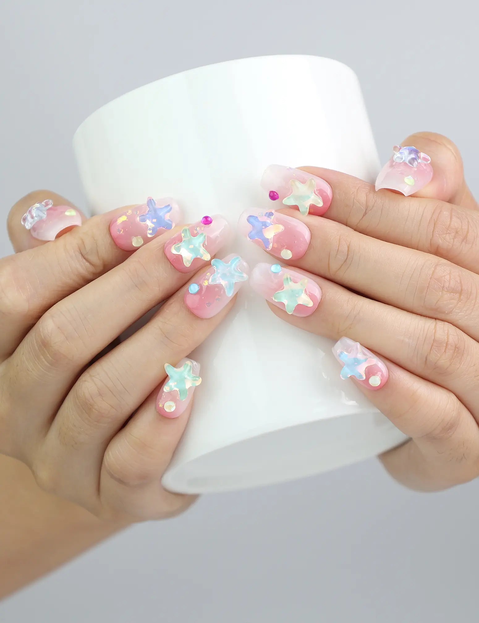 Hand holding a cup, displaying Naillover’s short pink coffin nails with sparkling base and colorful 3D sea star decor