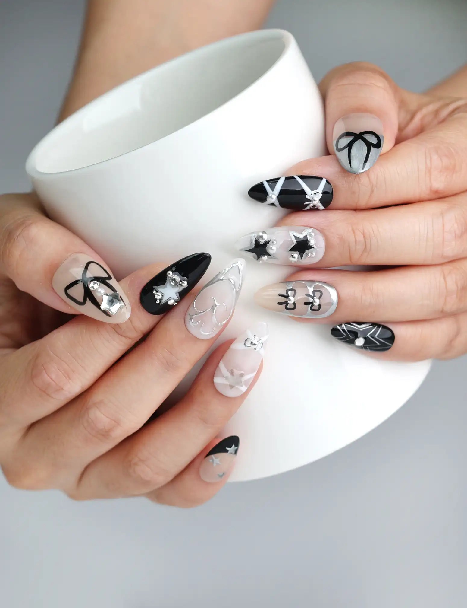 Hand holding a cup, showcasing Naillover’s nude and black press-on nails with glittery star accents and delicate bow embellishments