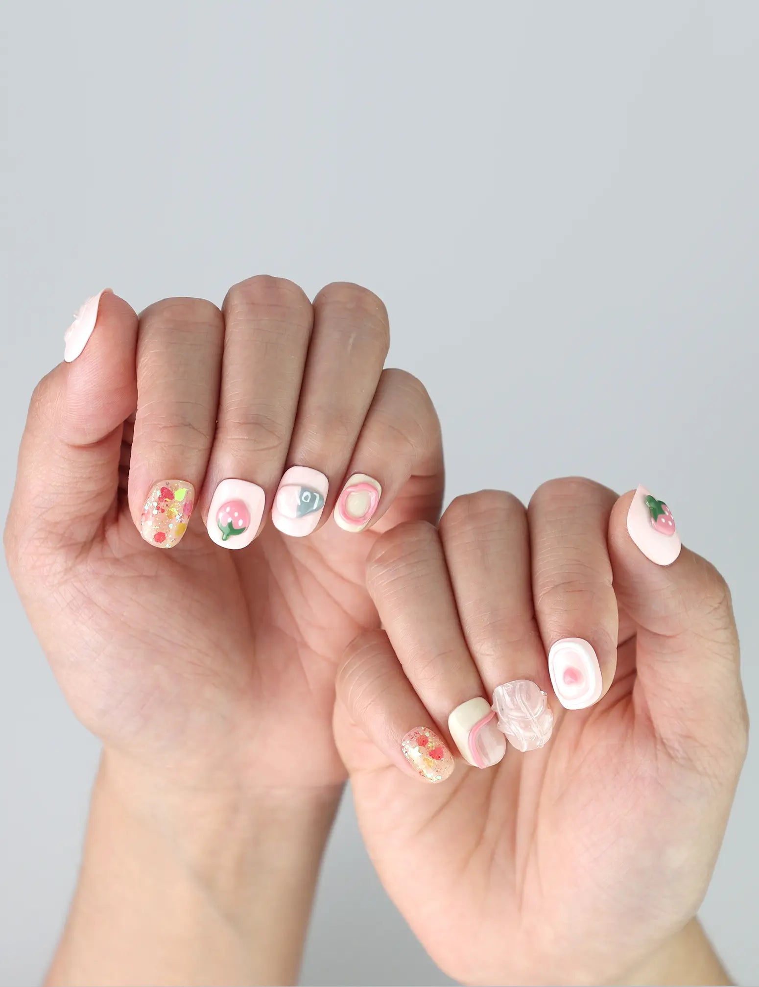 Delicate fingers posed to highlight short press on nails in pastel pink with charming strawberry and heart designs by Naillover